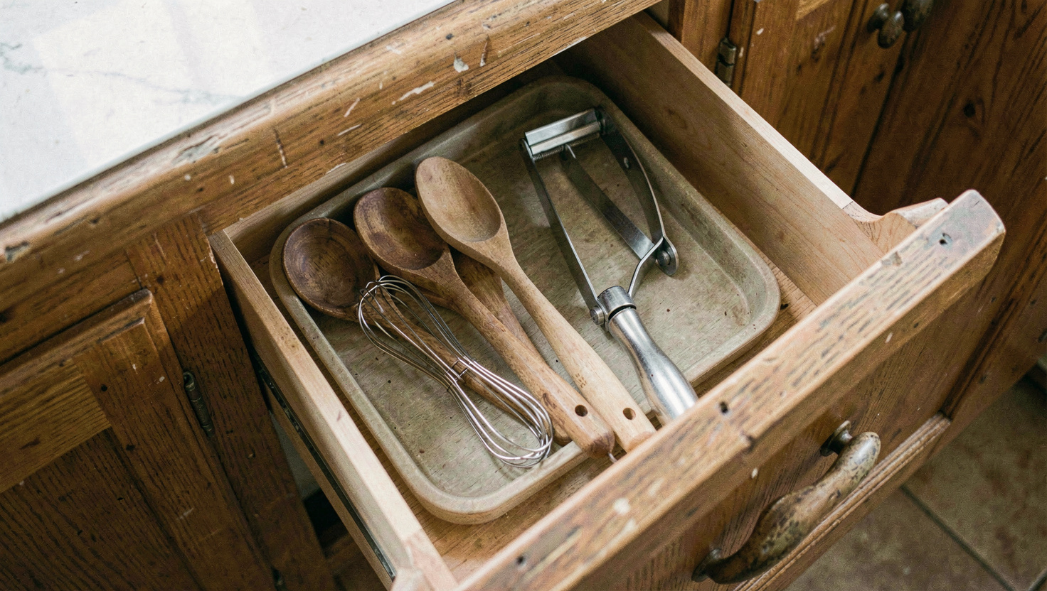 An open kitchen drawer with wooden spoons, a whisk and a peeler laid in a shallow tray, worn oak fronts around it, daylight from the left, photographed from above