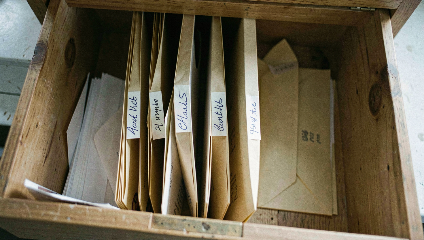 Four manila folders standing upright in a wooden drawer, hand lettered tabs reading in ink, a stack of envelopes beside them, cool north light, photographed from above