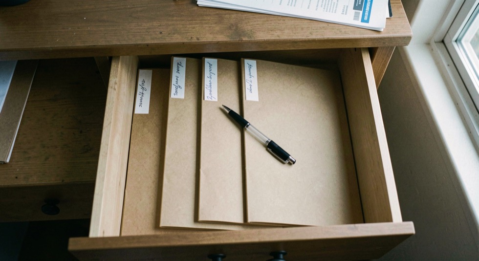 A wrapped framed picture lying flat on a wooden shelf in an interior cupboard, corners padded with card, bubble wrap bubbles facing outward, soft daylight from a doorway falling across the bundle.